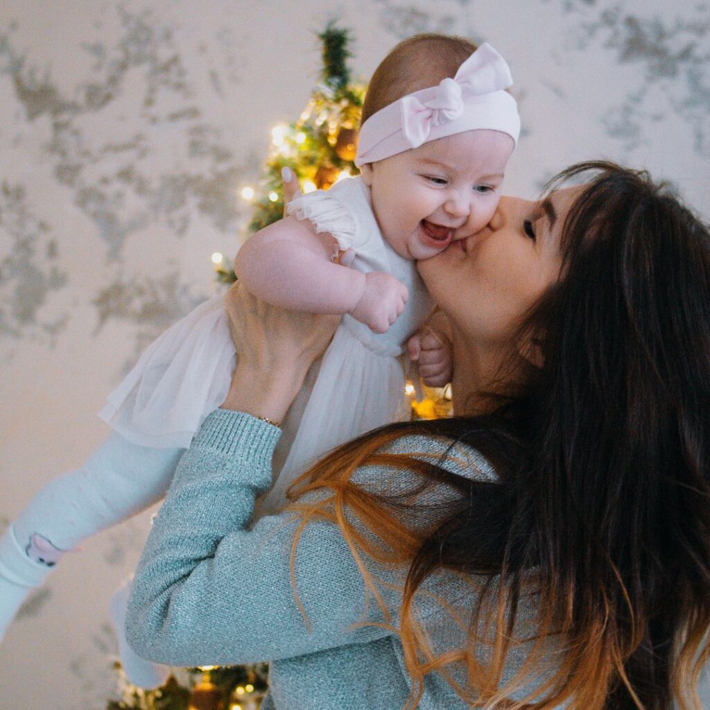Mom holds up baby and kisses baby on the cheek in front of a christmas tree